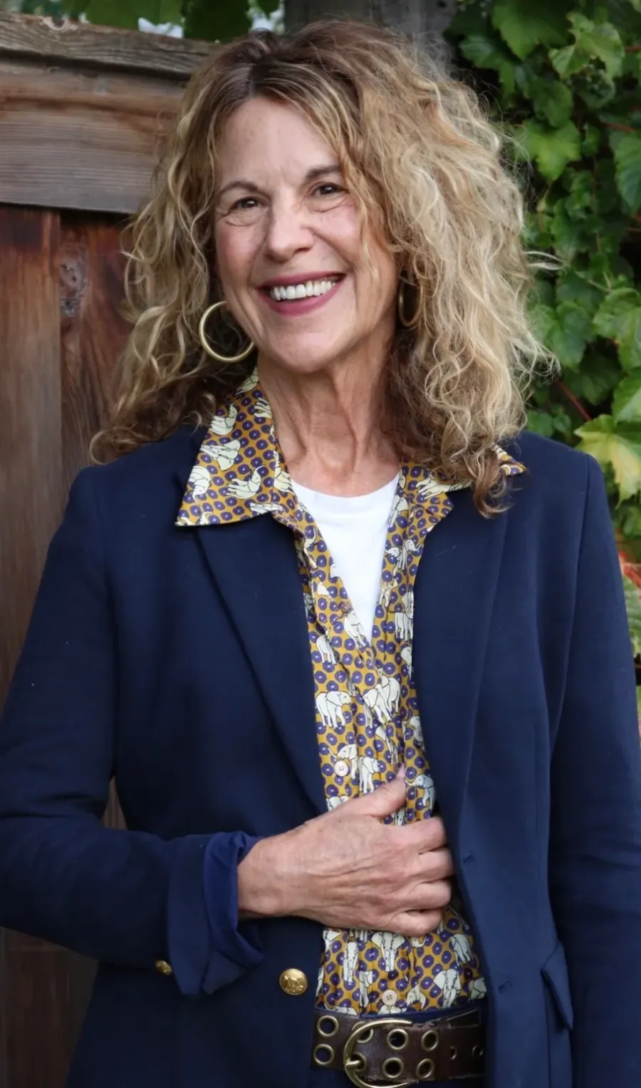 Sharon Davis smiling in a navy blazer, one hand at the heart, in front of a wooden gate and vines