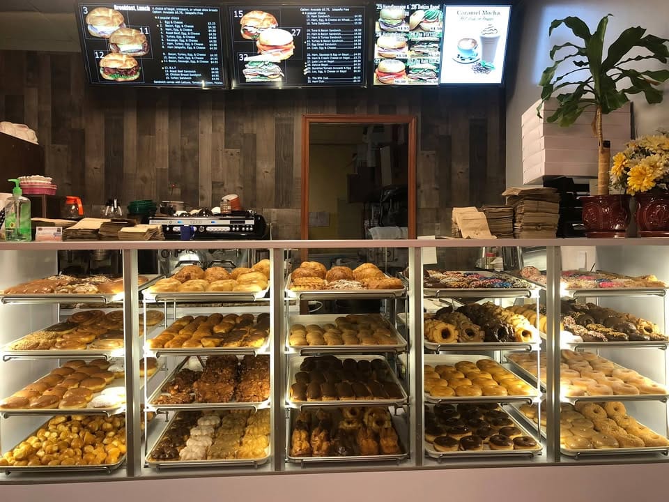 The full Sam's Donuts display case packed with trays of glazed, frosted, filled and sprinkled donuts under bright menu boards.